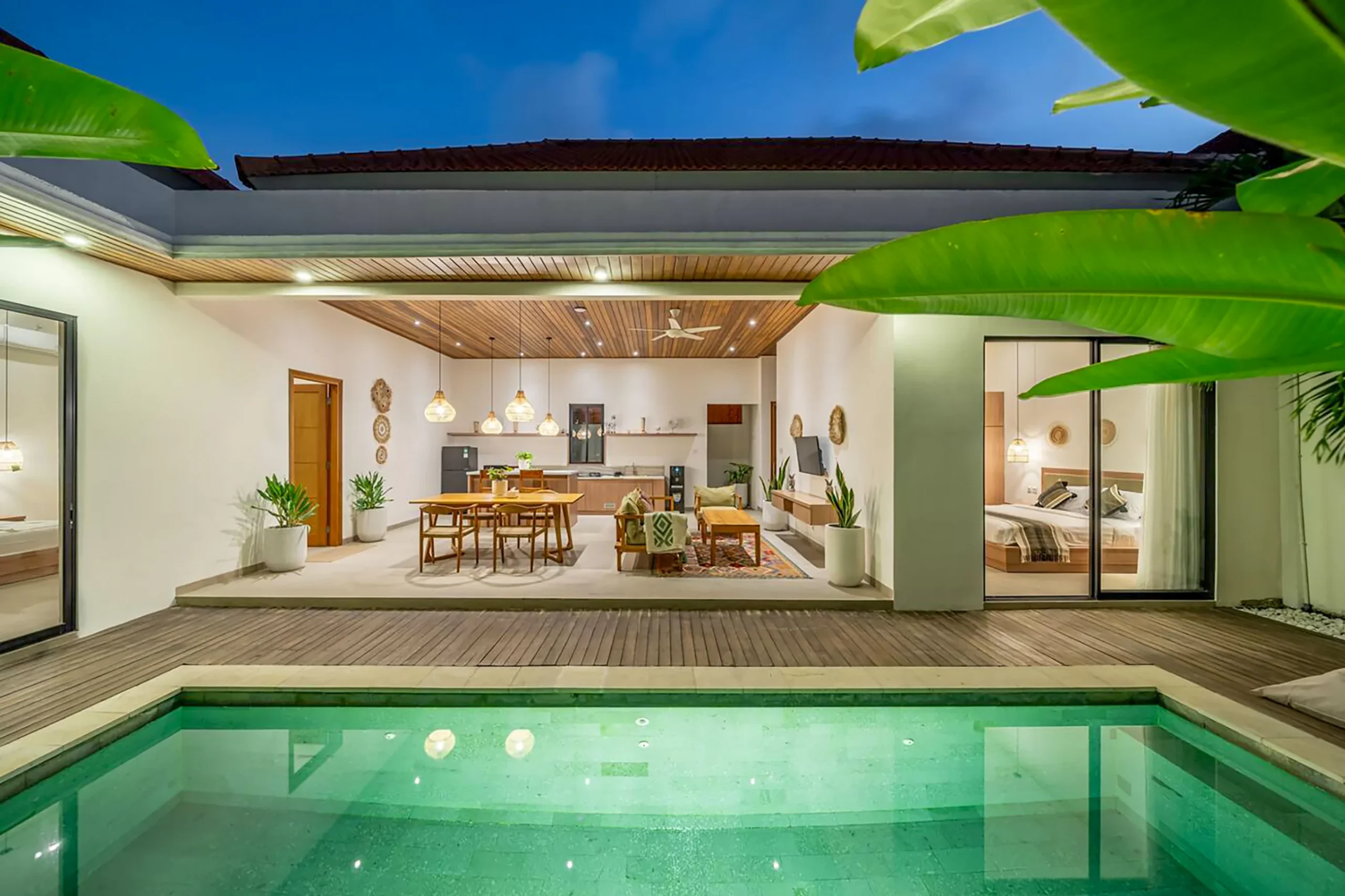 Villa LaBumbak Umalas Bali - illuminated private pool at blue hour with open-plan living, dining, rattan pendants and both bedrooms visible through glass, banana palms framing.