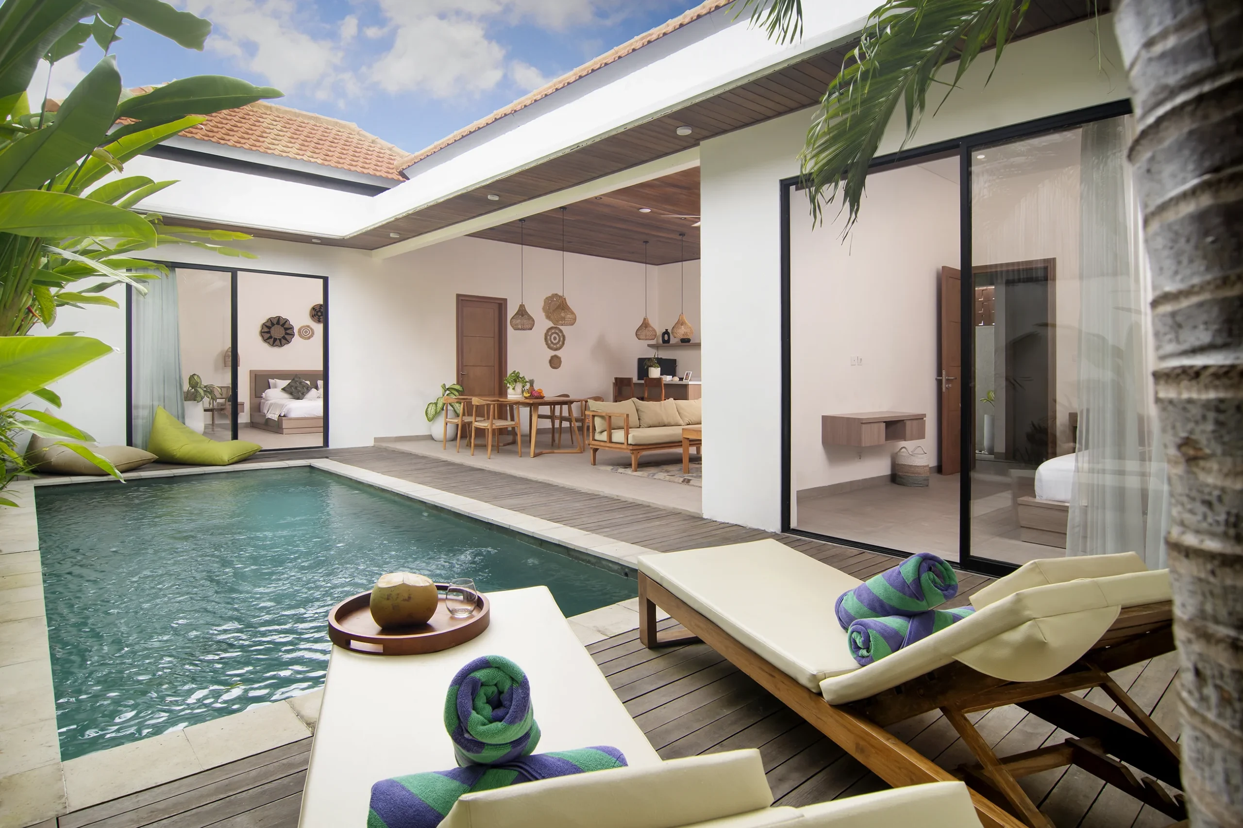 Villa LaBumbak Umalas Bali — daytime pool with two teak sunloungers, coconut tray, rolled towels, open-plan living room with rattan pendants behind, both bedrooms through glass, blue sky.