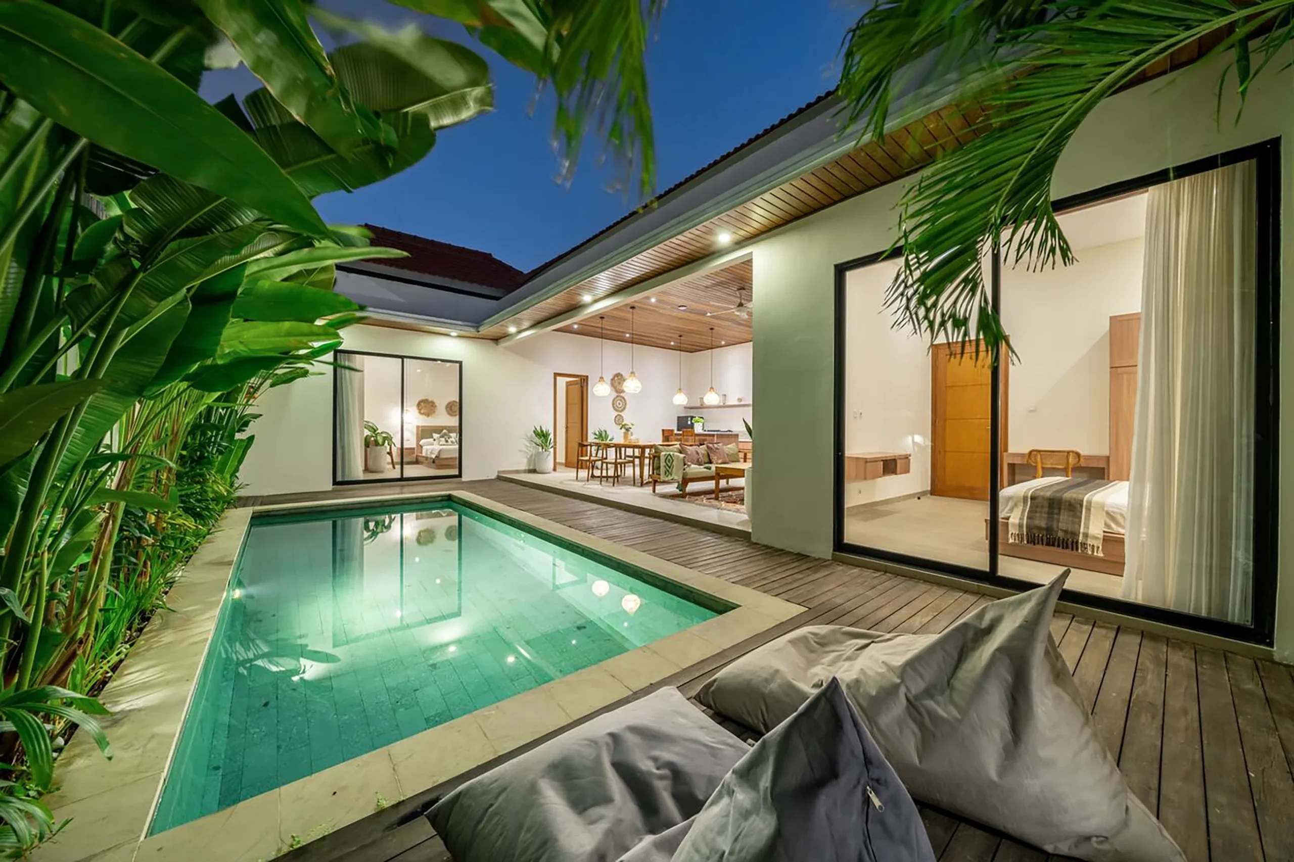 Villa LaBumbak Umalas Bali — illuminated private pool at twilight, grey bean bags on teak deck, open-plan living and both bedrooms glowing through glass, banana palms, blue dusk sky.