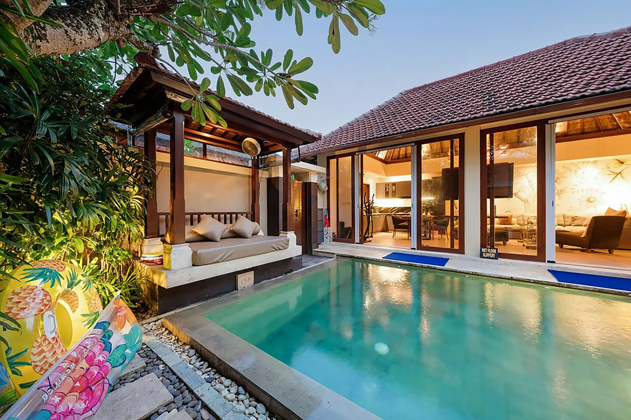 Private pool at twilight at Disini Villa 23, Seminyak Bali — illuminated pool with Balinese bale daybed pavilion, tropical garden, glass doors to glowing living room, terracotta roof, frangipani tree.