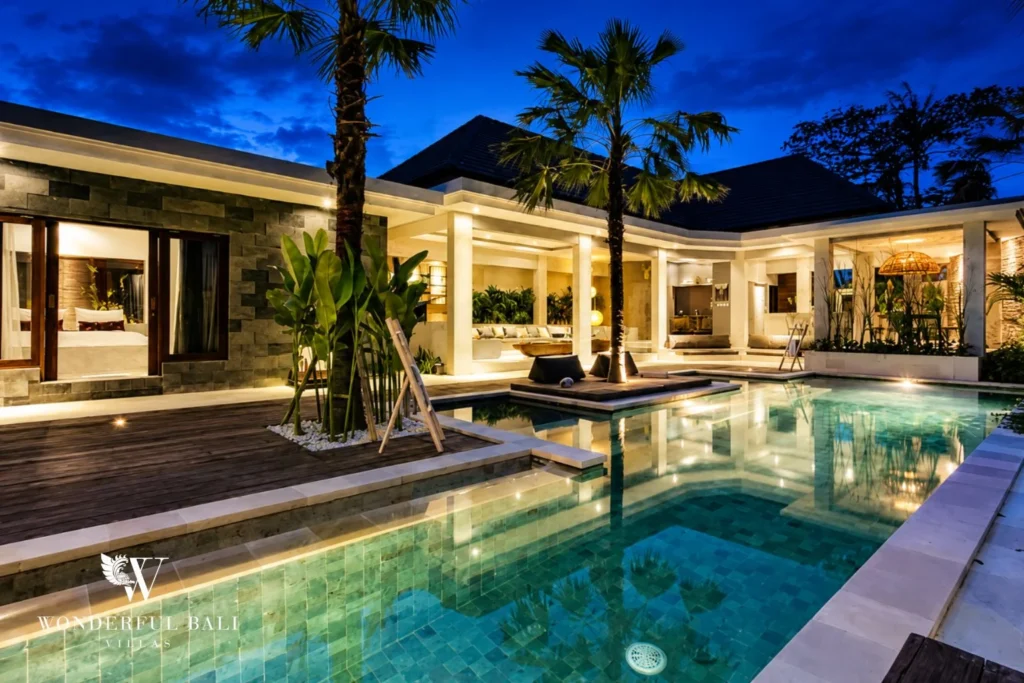Pool at evening with palms, lit pavilions and central sun-deck at villa in Seminyak, Bali