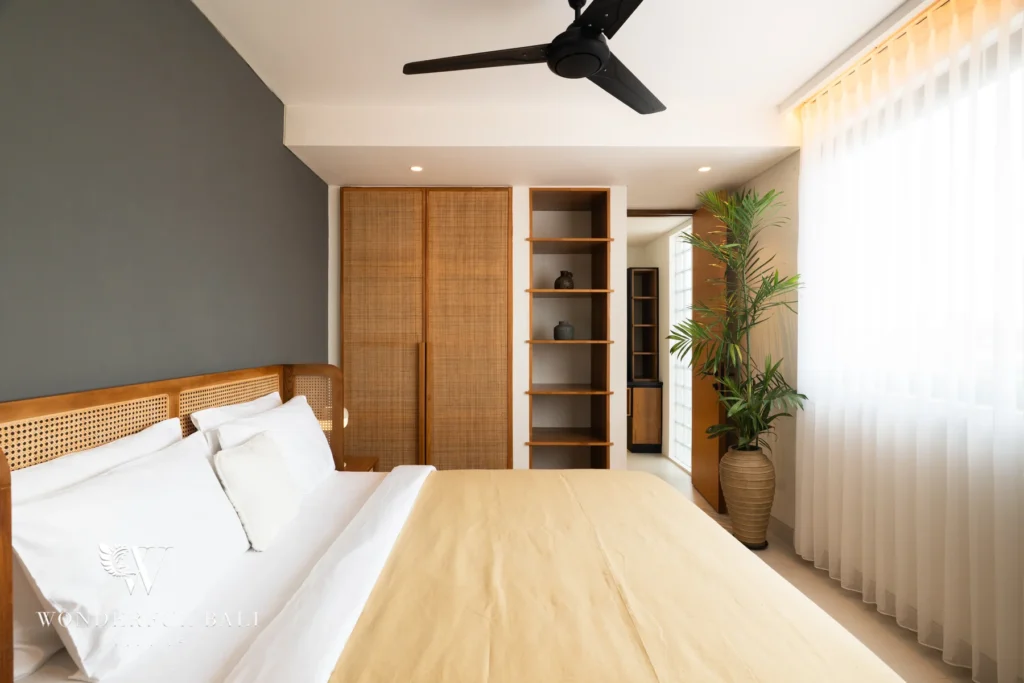 A bright en-suite bedroom with a cane headboard, grey feature wall, woven rattan wardrobe, open shelving and a potted palm beside sheer curtains at a 5-bedroom villa in Batu Bolong, Canggu, Bali