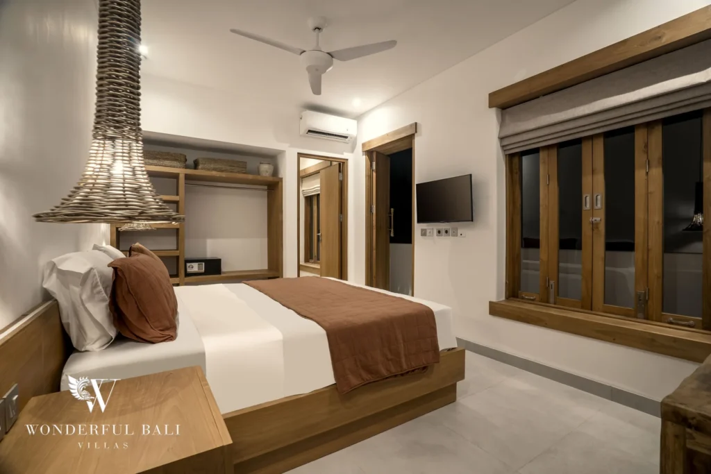 A king bed with a brown throw sits beneath a ceiling fan next to open wooden shelving, a wall-mounted TV and a wood-framed window at a 3-bedroom villa in Bingin, Uluwatu, Bali