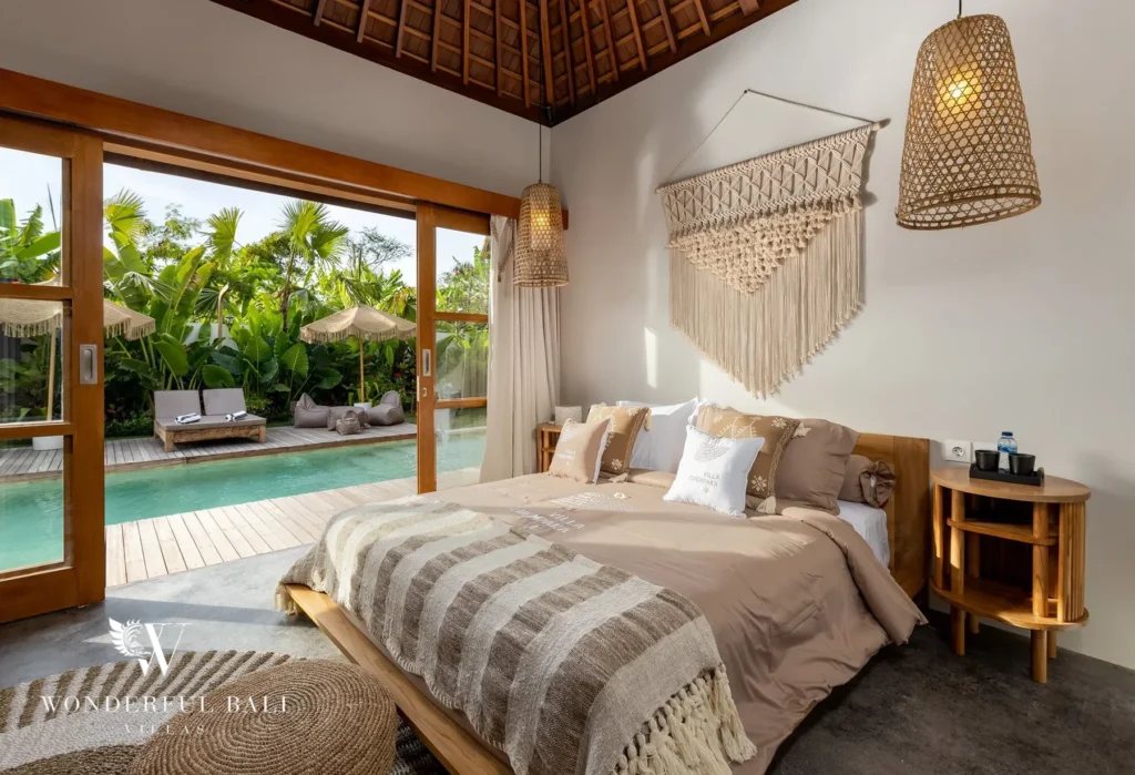 A boho bedroom with a king bed, macrame wall hanging and woven pendant lamps opens through sliding doors to a poolside lounge and tropical garden at a 4-bedroom villa in Canggu, Bali