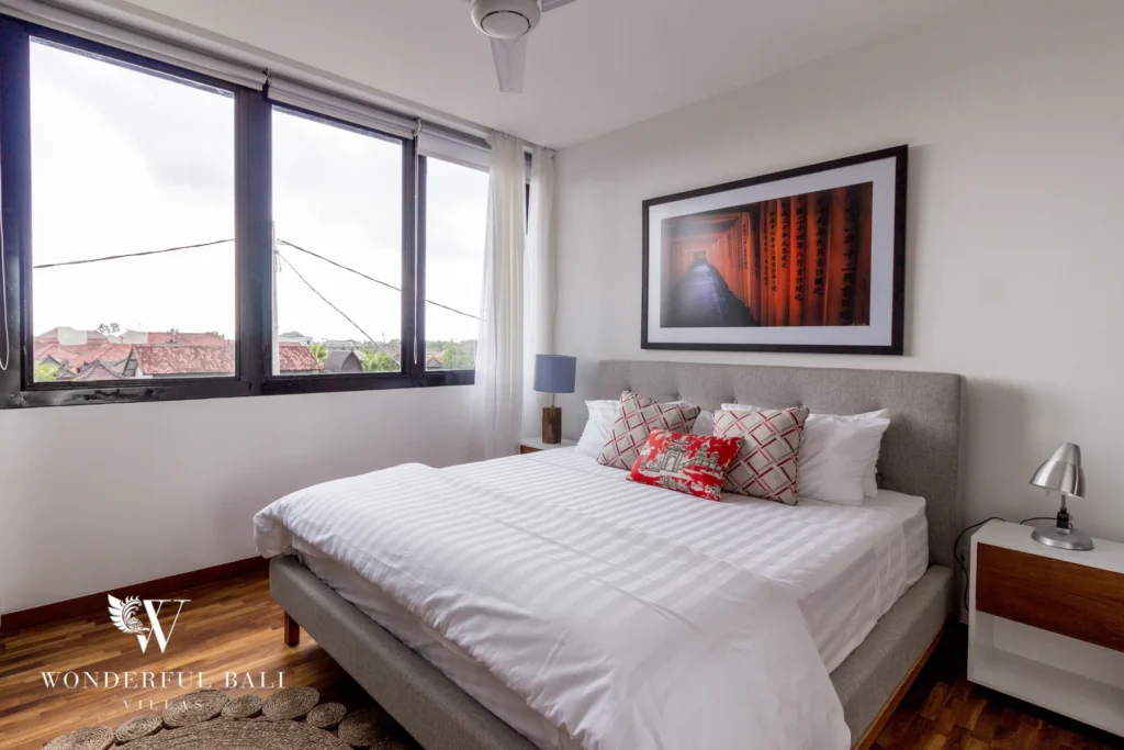 A bedroom with a grey upholstered bed, red torii-gate artwork and a corner window framing rooftop views over timber floors at a 4-bedroom villa in Batu Bolong, Canggu, Bali