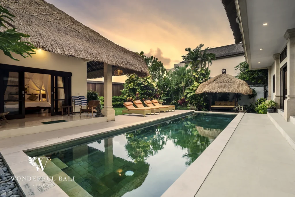A private swimming pool at dusk flanked by a thatched cabana, sun loungers and a pavilion bedroom with lush garden at a 4-bedroom villa in Seminyak, Bali