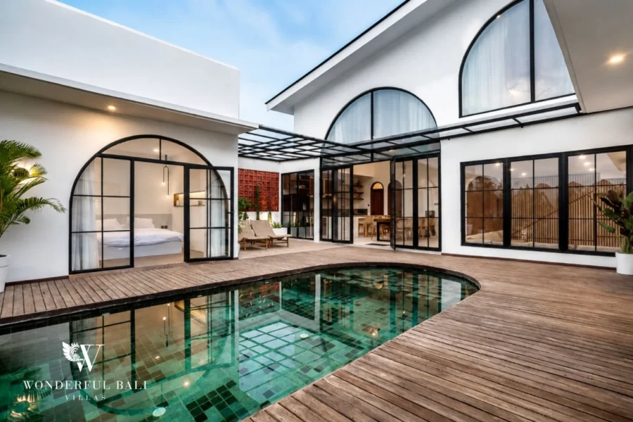 The green mosaic-tile pool and wooden-deck courtyard framed by black steel-frame arched glass doors opening to a bedroom at a 3-bedroom villa in Pererenan, Bali