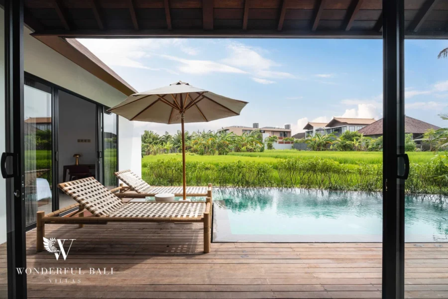 Two bamboo sun loungers under a canvas umbrella on a wooden deck beside a pool with open rice-field views at a 4-bedroom villa in Canggu, Bali