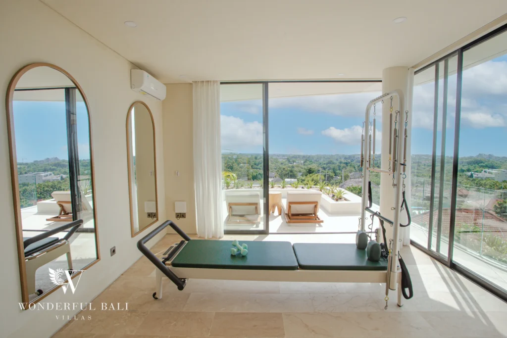 A home gym with a pilates reformer, arched mirrors and sliding glass doors opening to a terrace with sun loungers and a panoramic view at a 3-bedroom villa in Bingin, Uluwatu, Bali
