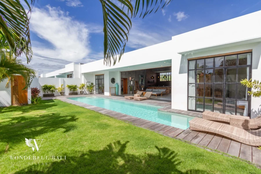 A white villa facade opens to a long private pool bordered by a timber sun deck and green lawn under a bright blue sky at a 3-bedroom villa in Seminyak, Bali