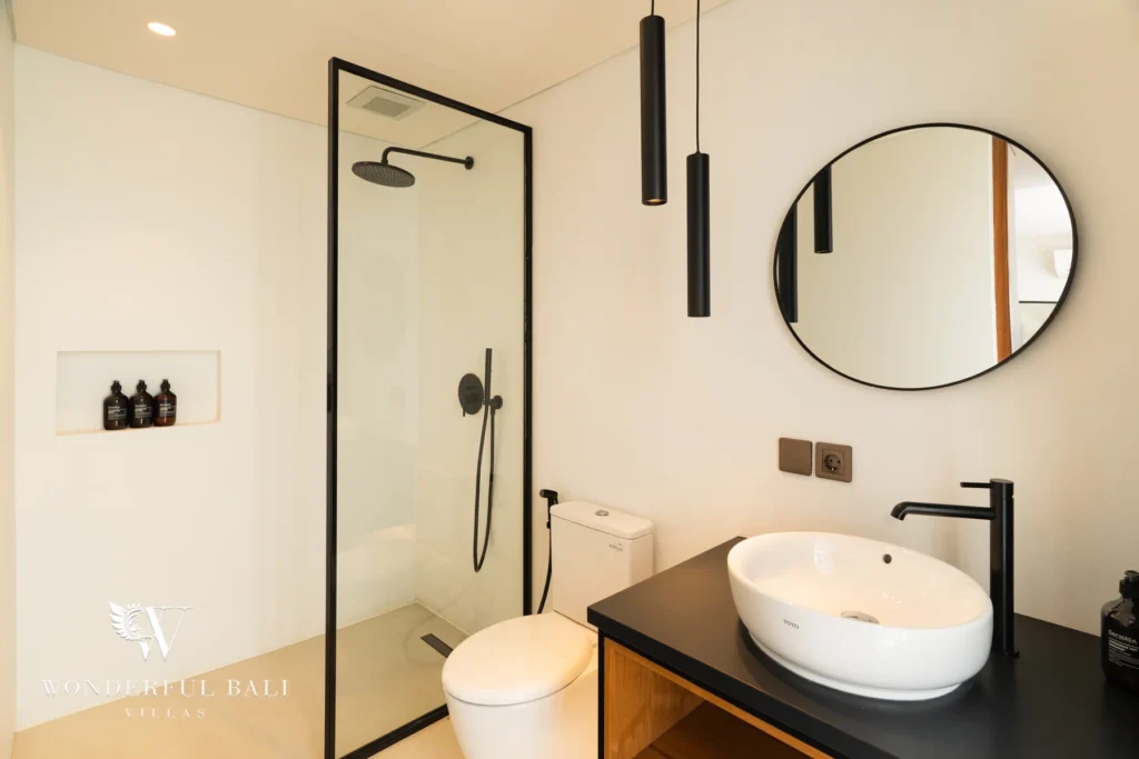 A minimalist en-suite bathroom with a glass walk-in rain shower, round black-framed mirror, vessel sink on a black timber vanity and cylindrical pendant lights at a 5-bedroom villa in Batu Bolong, Canggu, Bali
