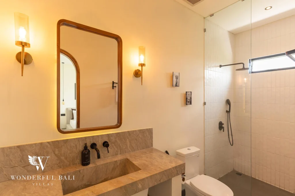 Warmly lit en-suite vanity with a stone sink, arched mirror and wall sconces beside a glass shower at a 2-bedroom villa in Canggu, Bali