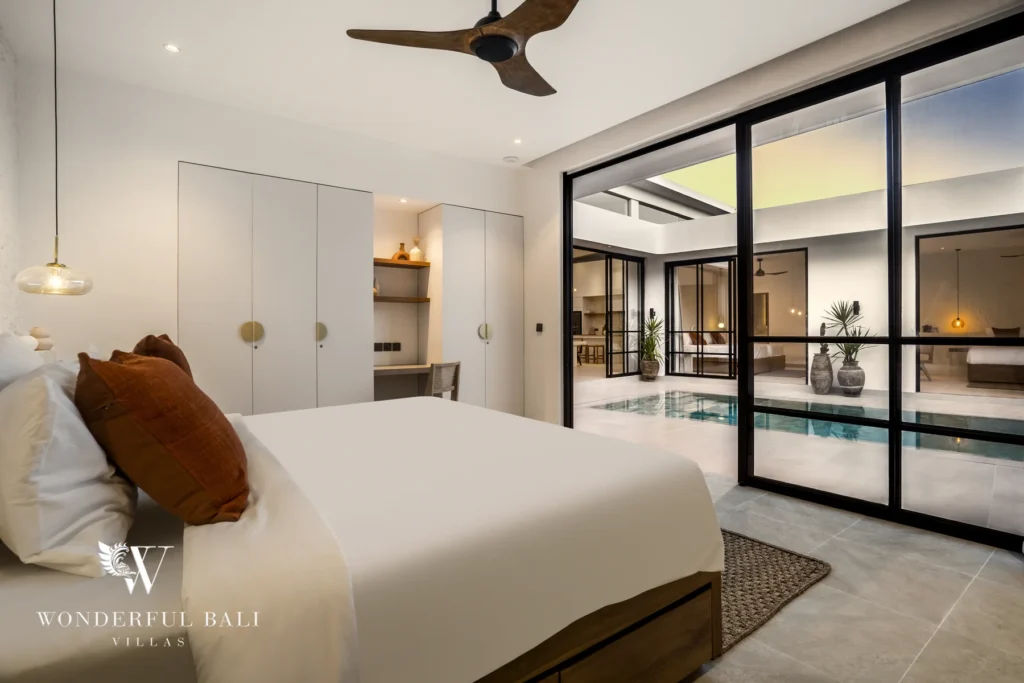 A bedroom with white bedding and rust cushions opening through wide glass sliding doors onto the pool courtyard at a 3-bedroom villa in Bingin, Uluwatu, Bali