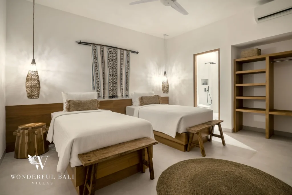 Two single beds with woven pendant lights, a patterned textile wall hanging and an en-suite shower and open shelving beyond at a 3-bedroom villa in Bingin, Uluwatu, Bali