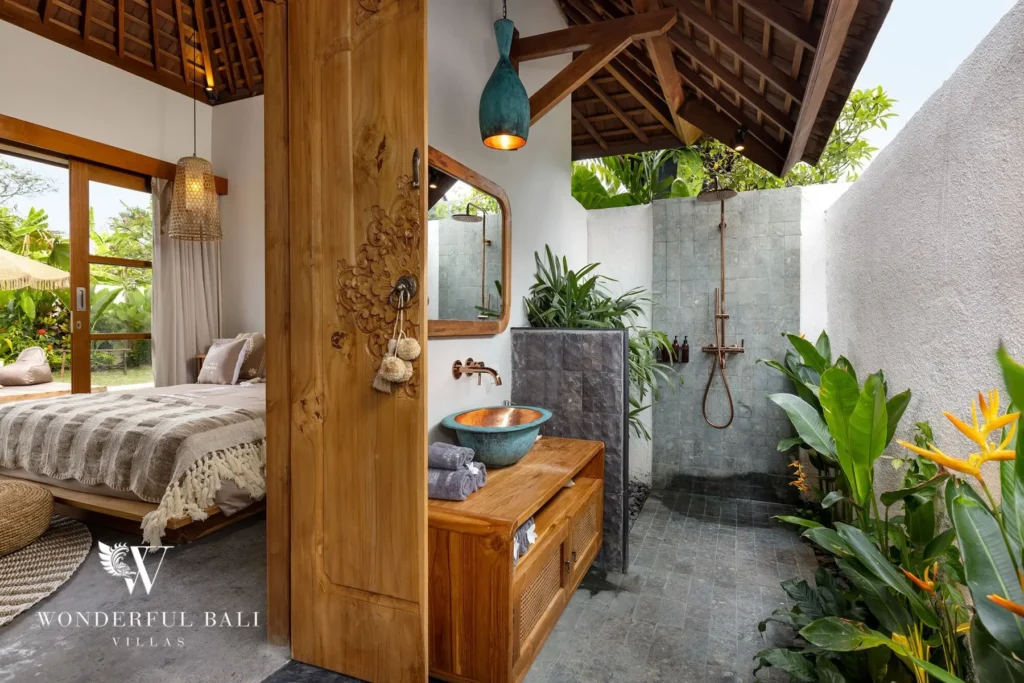 An open-air en-suite bathroom with a carved wood vanity, blue stone basin and copper shower sits among tropical plants beside a garden bungalow bedroom at a 4-bedroom villa in Canggu, Bali