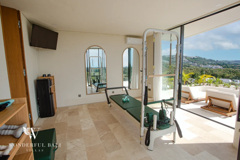 A private gym with reformer equipment, a wall-mounted TV and glass doors folded open to a sunlounger terrace overlooking green hills at a 3-bedroom villa in Bingin, Uluwatu, Bali