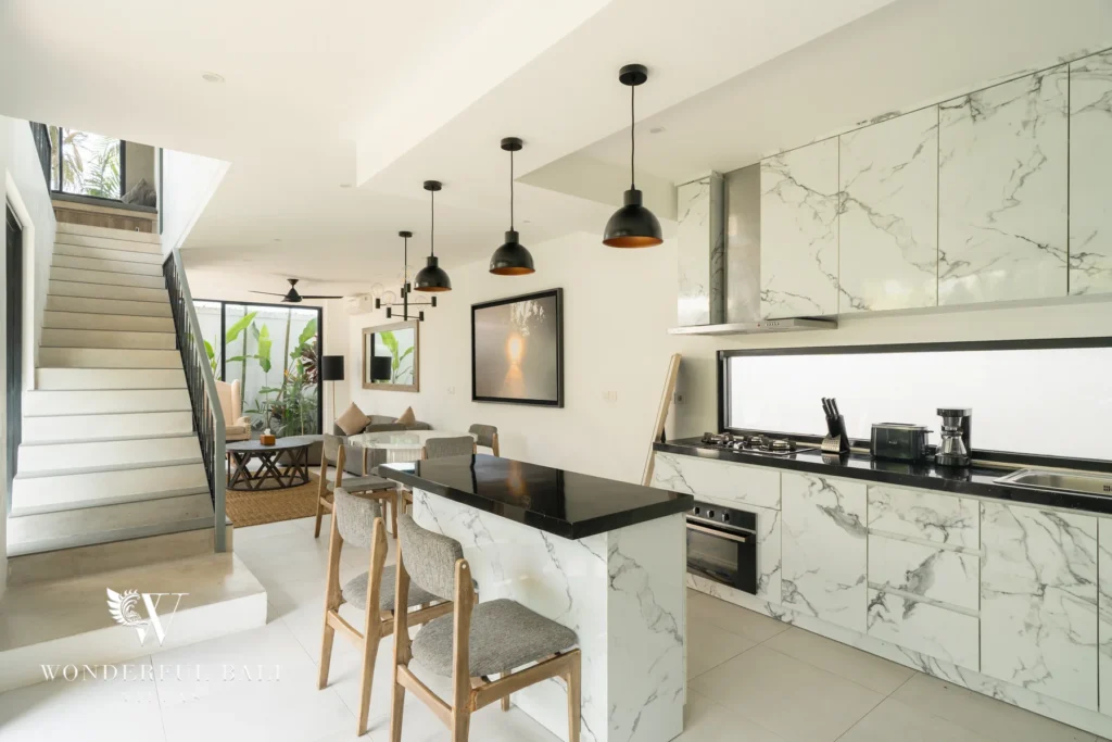 A white marble kitchen with a black-topped island bar, wooden stools, gas hob and pendant lights beside a staircase and lounge area at a 3-bedroom villa in Berawa, Bali