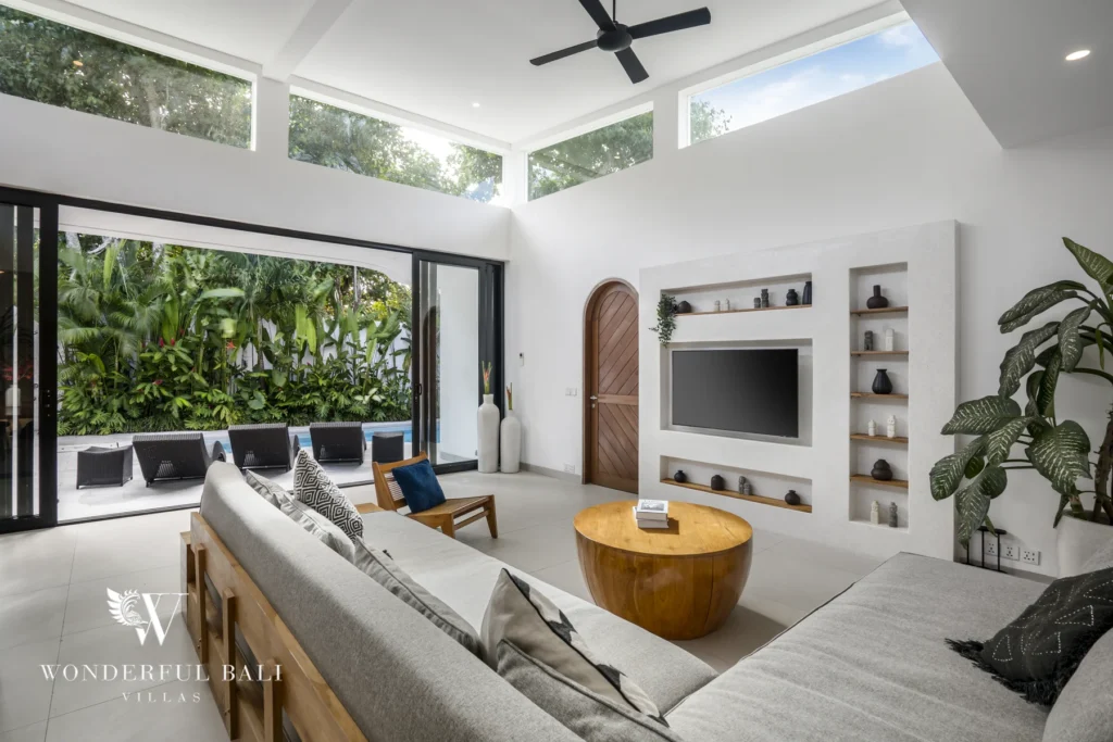 A bright open-plan living room with a grey sofa, wall-mounted TV and sliding glass doors opening to a leafy garden lounge at a 3-bedroom villa in Bingin, Uluwatu, Bali