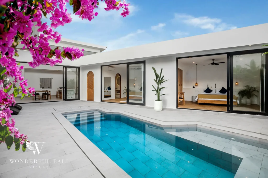 Bright daytime view across the turquoise pool toward floor-to-ceiling sliding glass doors opening into the bedrooms, framed by pink bougainvillea at a 3-bedroom villa in Bingin, Uluwatu, Bali
