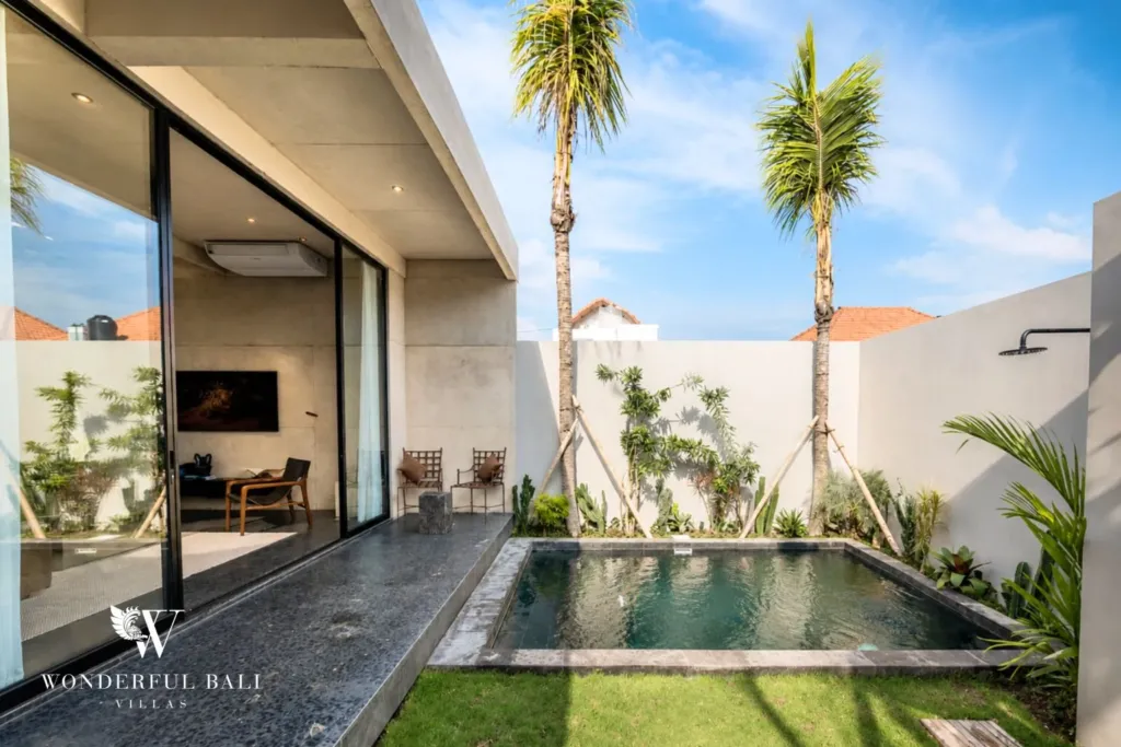 A private plunge pool bordered by lawn and palm trees with an outdoor shower and glass doors opening to the living room at a 2-bedroom villa in Seseh, Bali