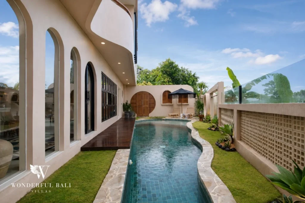 A long turquoise pool runs alongside the curved villa wall and wooden deck bordered by lawn at a 3-bedroom villa in Pererenan, Bali