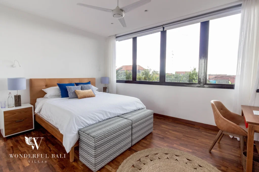 A bedroom with a timber-framed bed, blue and tan cushions, a large corner window and a wooden desk with chair at a 4-bedroom villa in Batu Bolong, Canggu, Bali