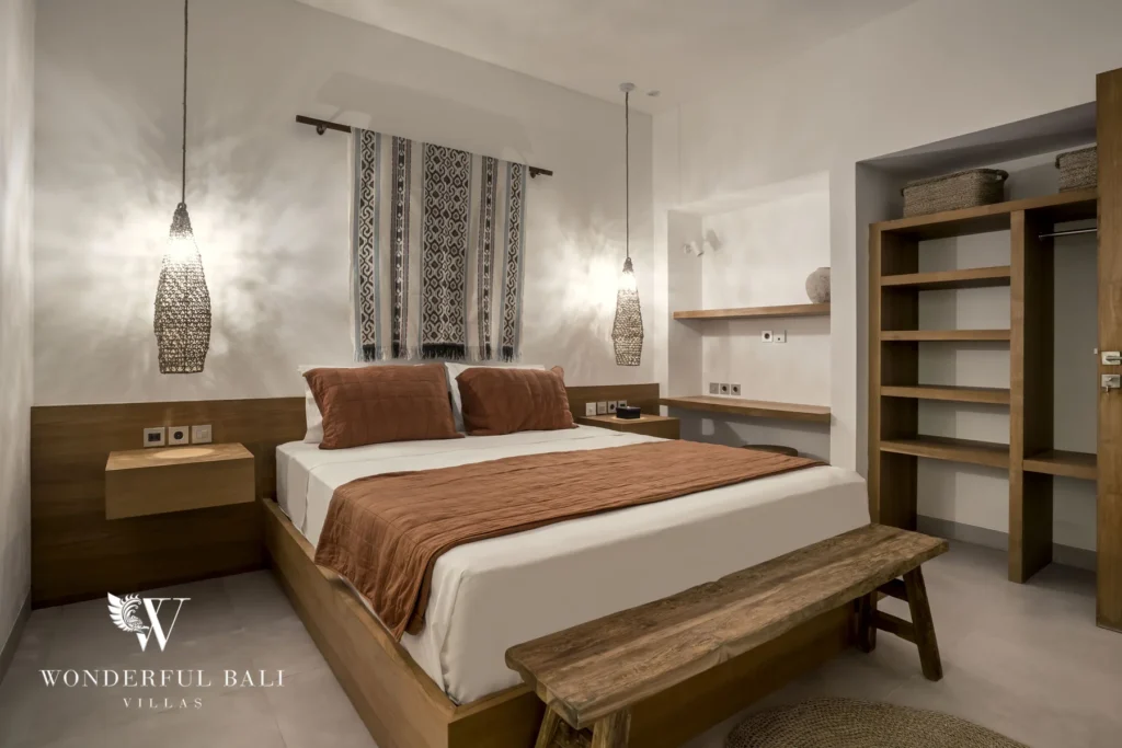 A king bed with a rust-colored throw framed by pendant lamps and a woven textile hanging, with a wooden bench and open shelving alongside at a 3-bedroom villa in Bingin, Uluwatu, Bali