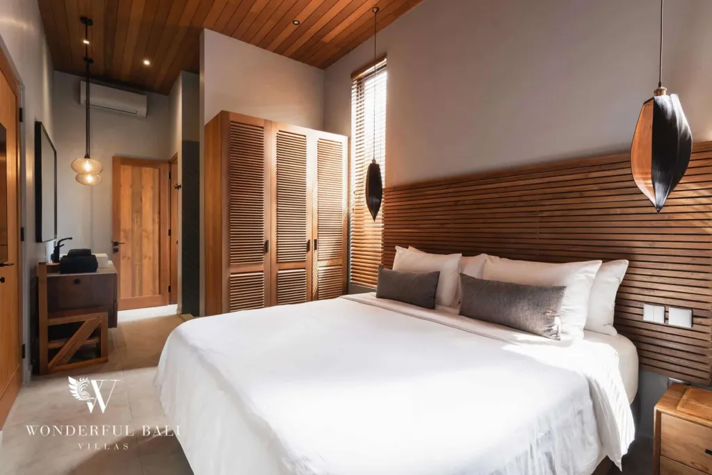 2BR villa for rent in Pererenan — bedroom, a bedroom with a wood-slat headboard, louvered timber wardrobe, plank ceiling and pendant lights beside a bright window at a 2-bedroom villa