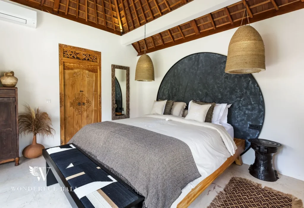 A bedroom with a king bed, dark rounded headboard, carved wooden door and woven pendant lights under a high timber ceiling at a 4-bedroom villa in Canggu, Bali