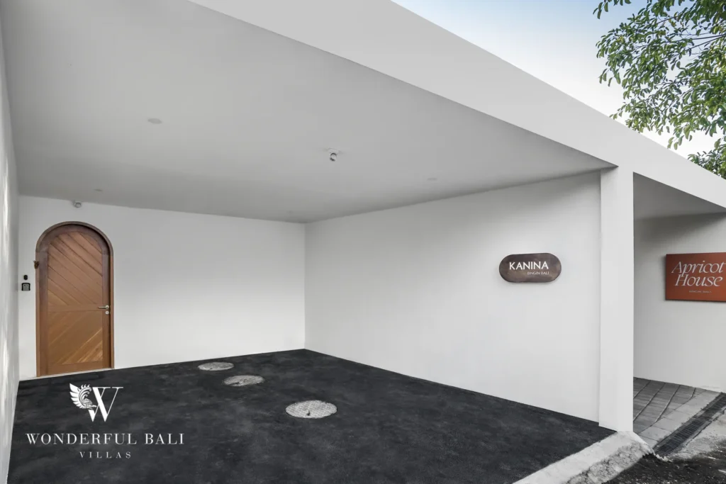 Minimalist white entrance area with an arched wooden door and a signboard beside a covered parking space at a 3-bedroom villa in Bingin, Uluwatu, Bali