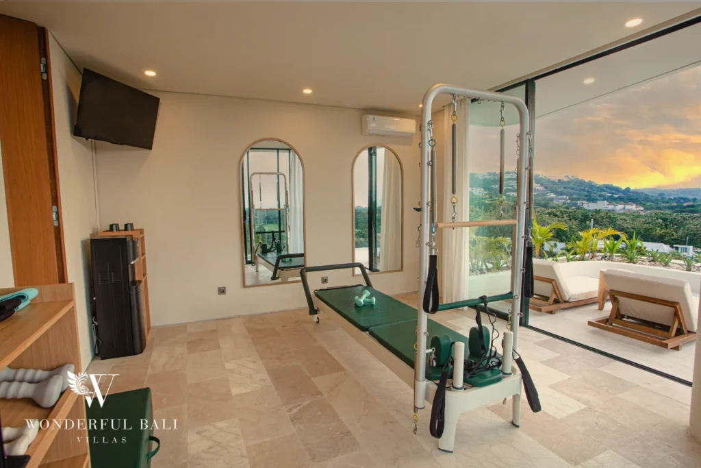 A gym with pilates equipment and arched mirrors opening onto a terrace with a golden sunset over distant hills at a 3-bedroom villa in Bingin, Uluwatu, Bali