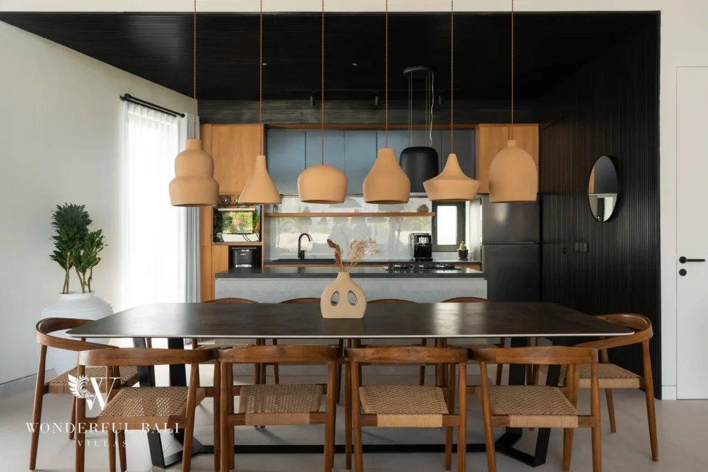 An open dining area with a long dark table, woven timber chairs and terracotta pendant lights in front of a modern kitchen with black slatted walls at a 5-bedroom villa in Batu Bolong, Canggu, Bali