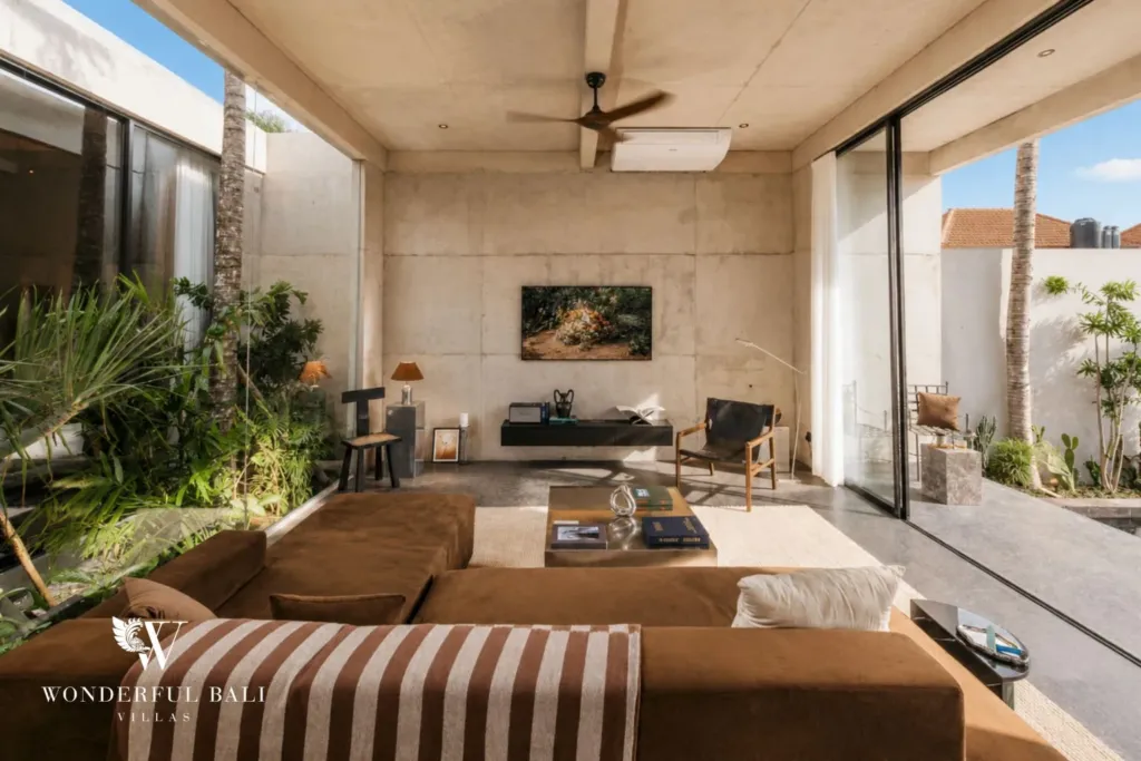 Open living room with a brown sofa, striped daybed, ceiling fan and exposed concrete walls framed by full-height glass doors and greenery at a 2-bedroom villa in Seseh, Bali