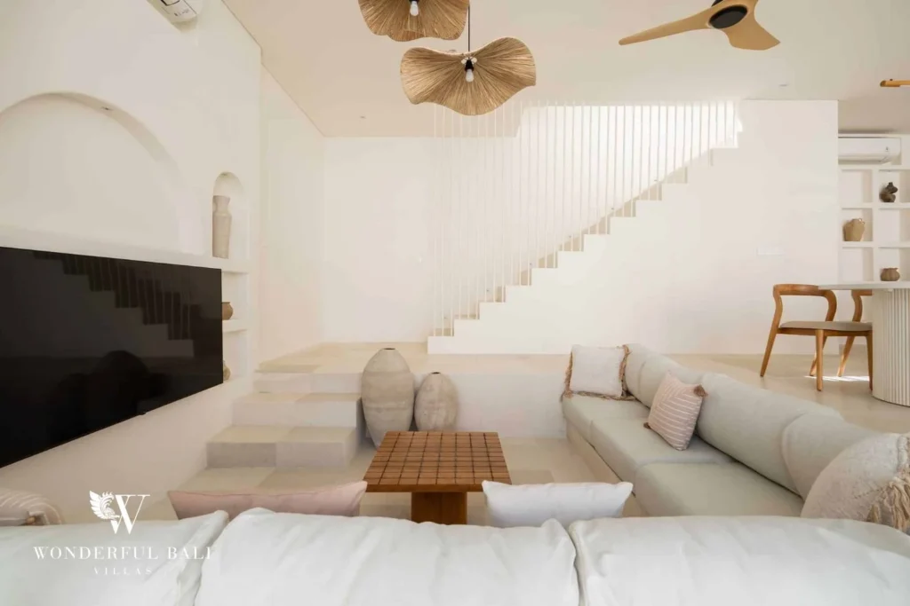 An open lounge with a low sectional sofa, a wall-mounted TV, woven pendant lights and a pale staircase, at a 2-bedroom villa in Bingin, Uluwatu, Bali