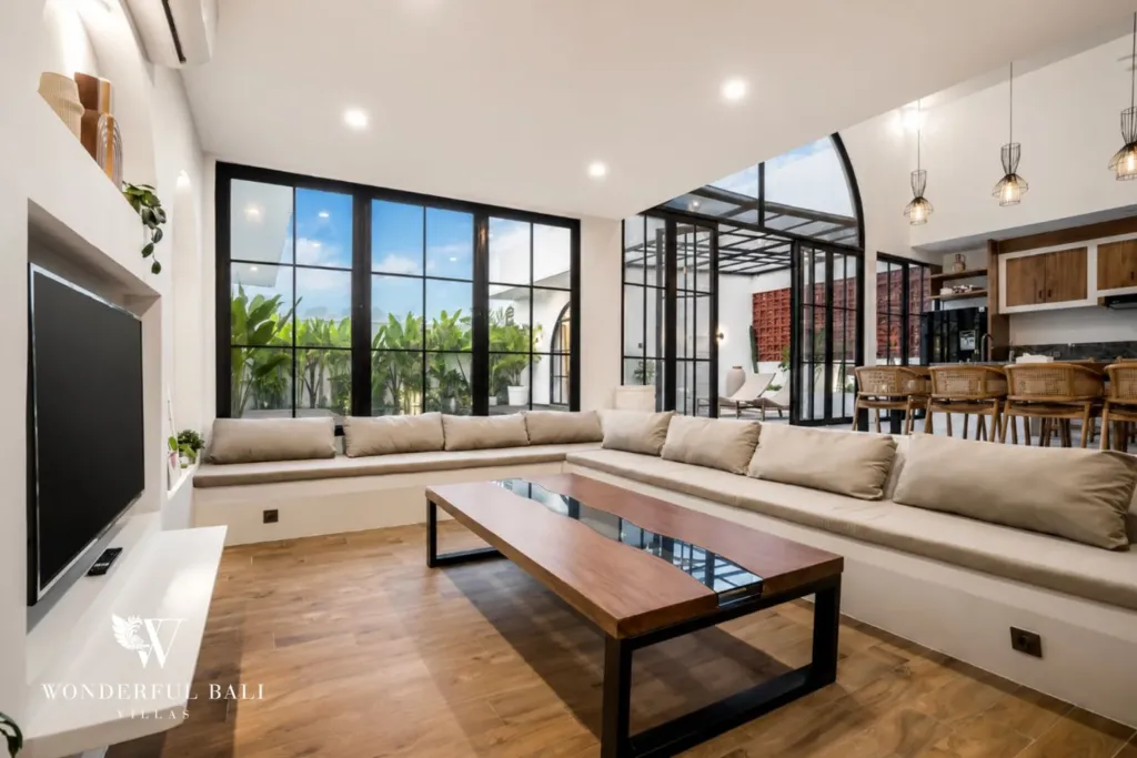 A bright open-plan living room with a beige L-shaped sofa, wooden coffee table, and tall black steel-frame windows at a 3-bedroom villa in Pererenan, Bali