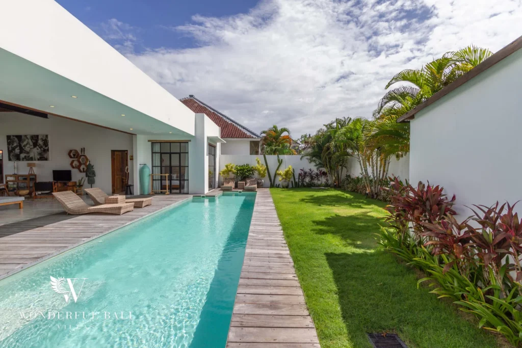 A slim lap pool runs alongside a timber walkway and manicured lawn with tropical palms and shrubs beside the villa at a 3-bedroom villa in Seminyak, Bali
