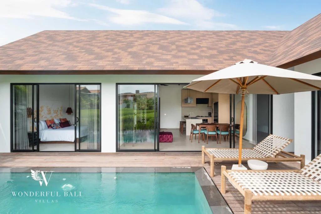 The tiled-roof villa facade with large glass sliding doors opening onto a bedroom and open-plan kitchen-dining area beside the pool and loungers at a 4-bedroom villa in Canggu, Bali