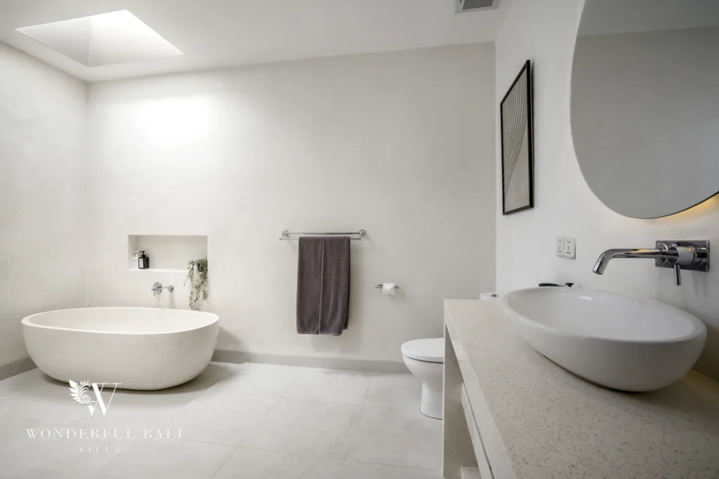 A minimalist white bathroom with a freestanding stone bathtub, twin vessel basin and round mirror at a 3-bedroom villa in Bingin, Uluwatu, Bali
