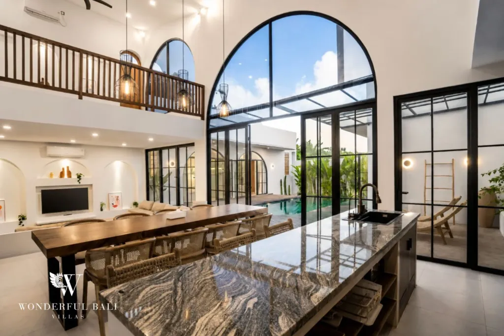 A soaring open-plan dining and kitchen area with a marble island beneath a towering arched window and a wooden mezzanine loft at a 3-bedroom villa in Pererenan, Bali