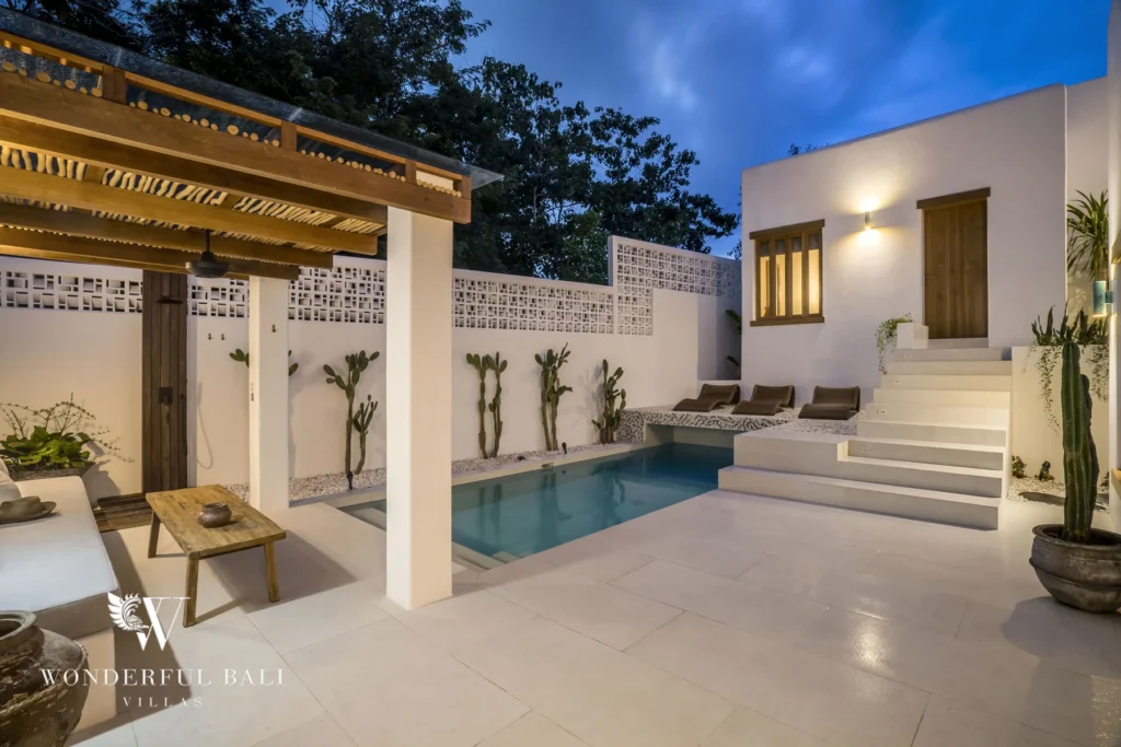 A private plunge pool with a covered lounge and wooden table glowing at dusk beside a white staircase and decorative breeze-block walls at a 3-bedroom villa in Bingin, Uluwatu, Bali