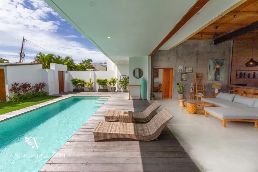 Rattan sun loungers rest on a timber deck beside the pool next to a concrete-walled lounge with a large painting at a 3-bedroom villa in Seminyak, Bali