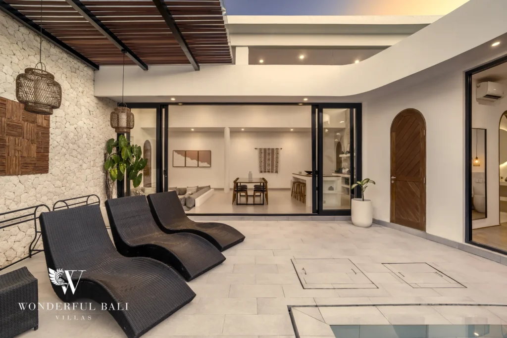 Black wicker sun loungers on the poolside terrace beside a stone accent wall and rattan lanterns at dusk at a 3-bedroom villa in Bingin, Uluwatu, Bali