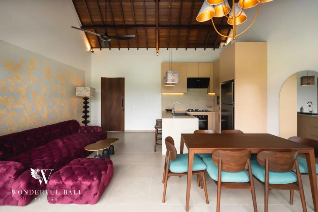 A high vaulted timber-beamed living space with a tufted magenta velvet sofa, a wooden dining table with teal chairs and an open kitchen behind at a 4-bedroom villa in Canggu, Bali