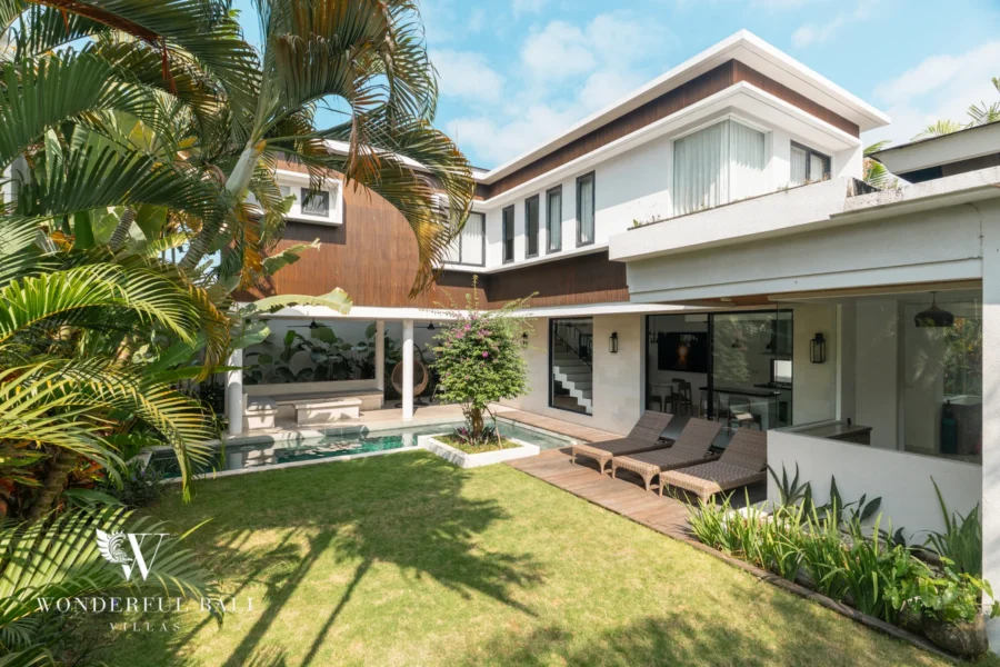 A garden view of the white and timber villa exterior with pool, wooden deck, sun loungers and covered outdoor seating at a 3-bedroom villa in Berawa, Bali