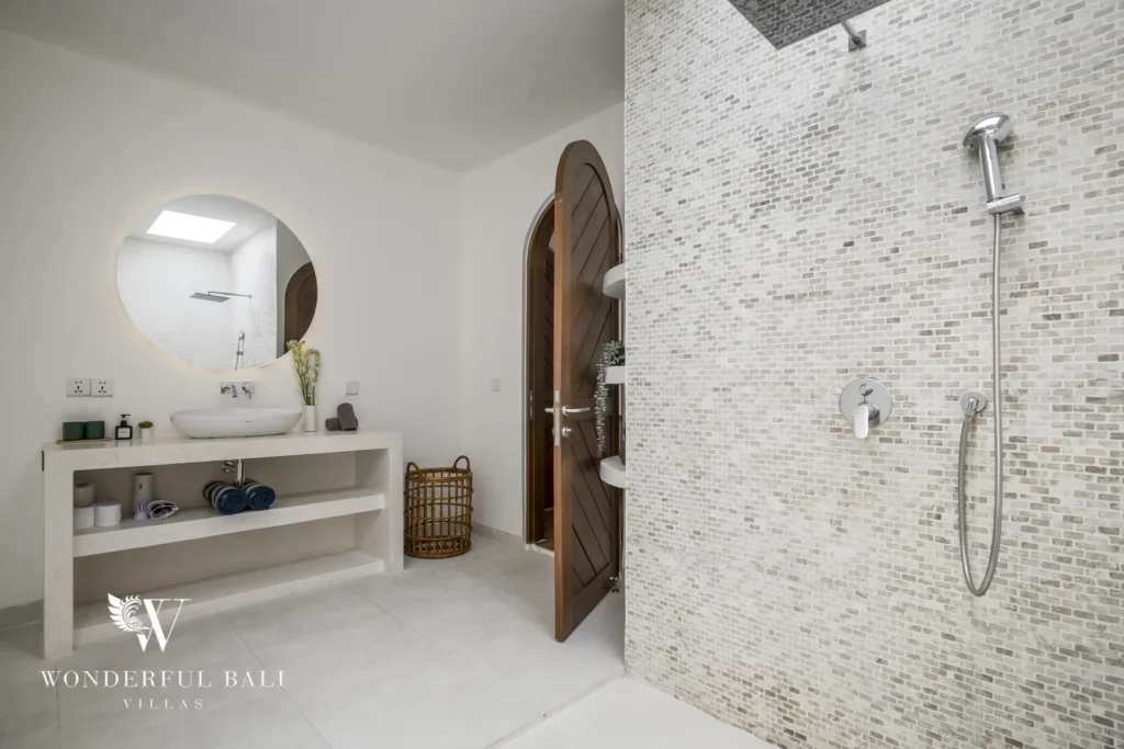 A bathroom with a mosaic-tiled walk-in shower, stone vanity, round mirror and arched wooden door at a 3-bedroom villa in Bingin, Uluwatu, Bali