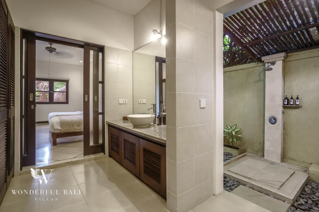 A bathroom with vanity and mirror leading to a semi-open rain shower with pebble stones adjoining a bedroom at a 4-bedroom villa in Seminyak, Bali