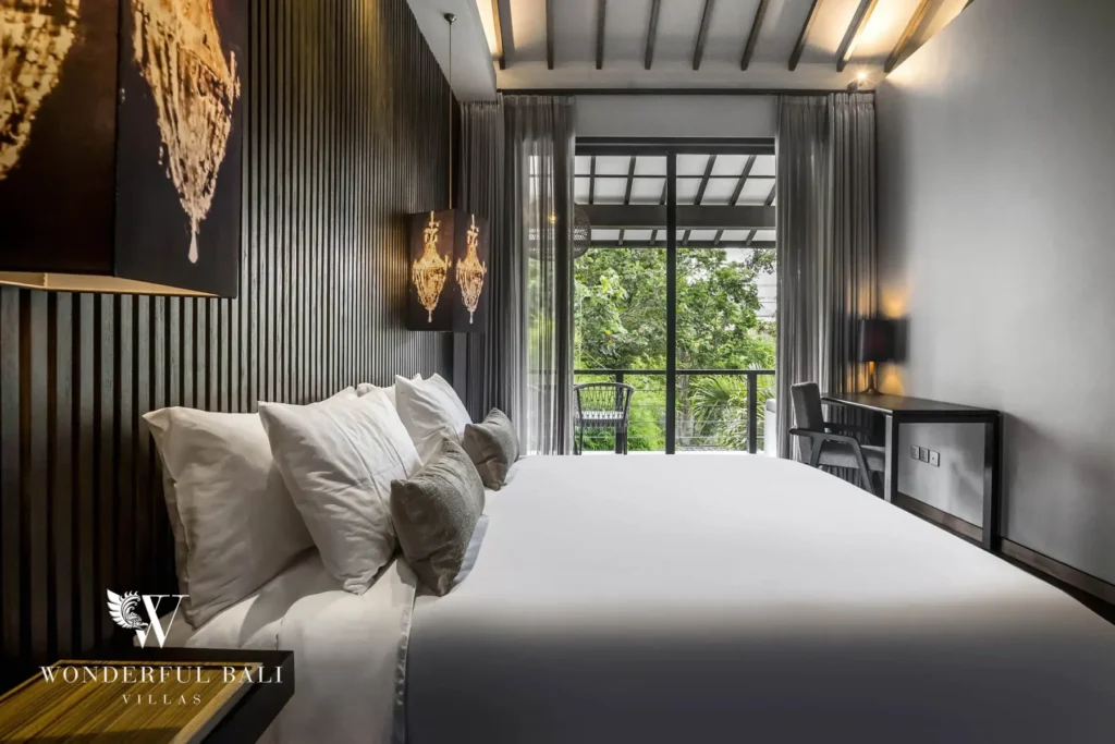 A side view of a bedroom with a cushioned white bed, a workspace desk and glass doors leading to a balcony with greenery beyond at a 3-bedroom villa in Pererenan, Bali