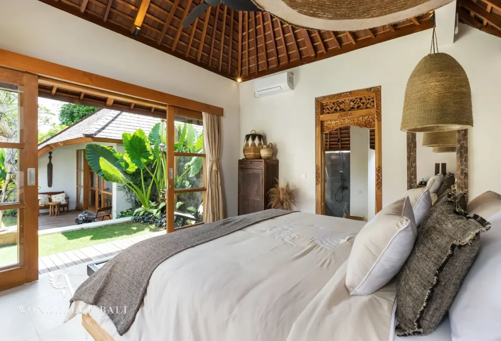A bright bedroom with a king bed and woven pendant lamp opens through sliding glass doors onto a decked garden with lush greenery at a 4-bedroom villa in Canggu, Bali