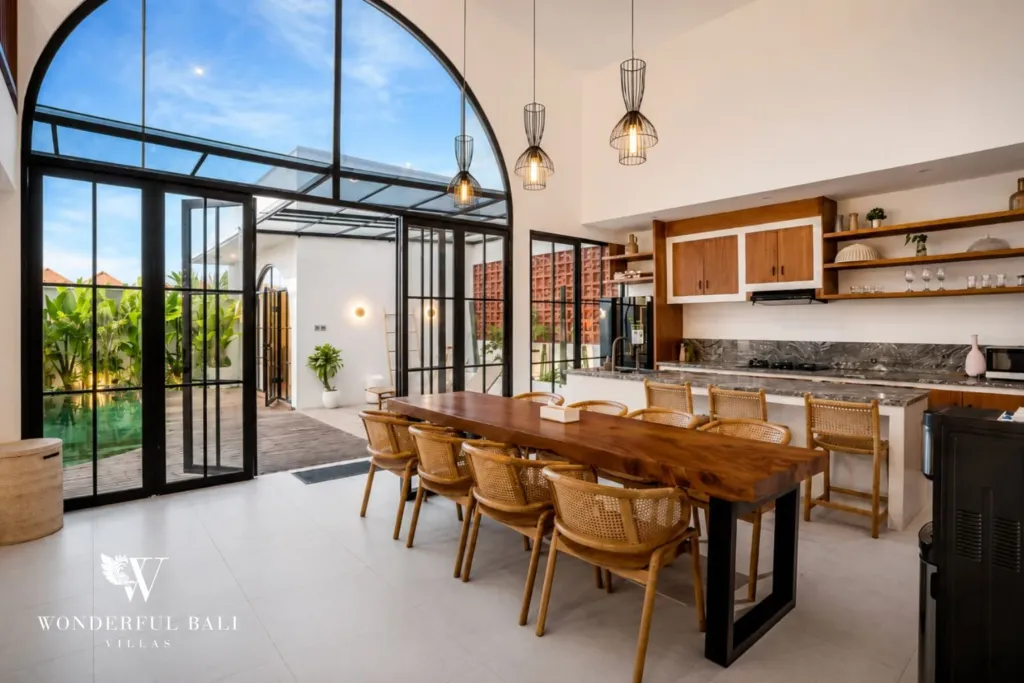 A live-edge wooden dining table and wood-cabinet kitchen sit under a dramatic double-height arched window at a 3-bedroom villa in Pererenan, Bali
