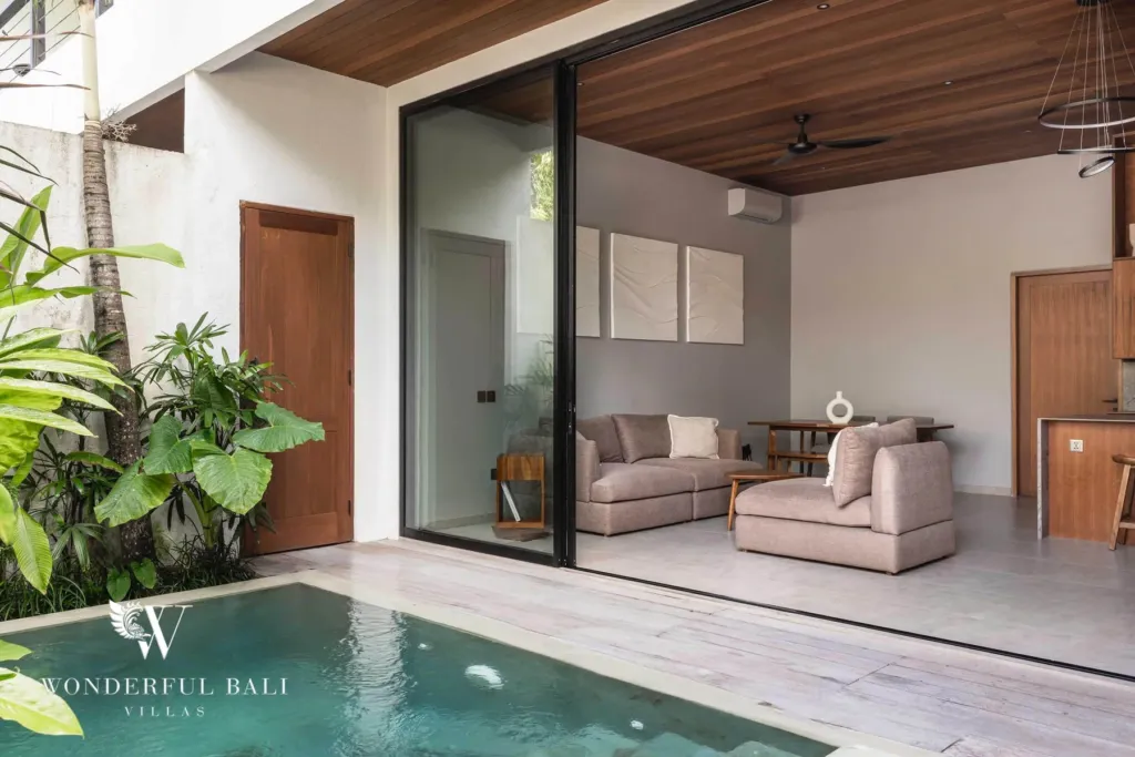 2BR villa for rent in Pererenan — living room, a sliding glass wall opening a linen-sofa lounge onto a timber deck and private plunge pool framed by tropical planting at a 2-bedroom villa